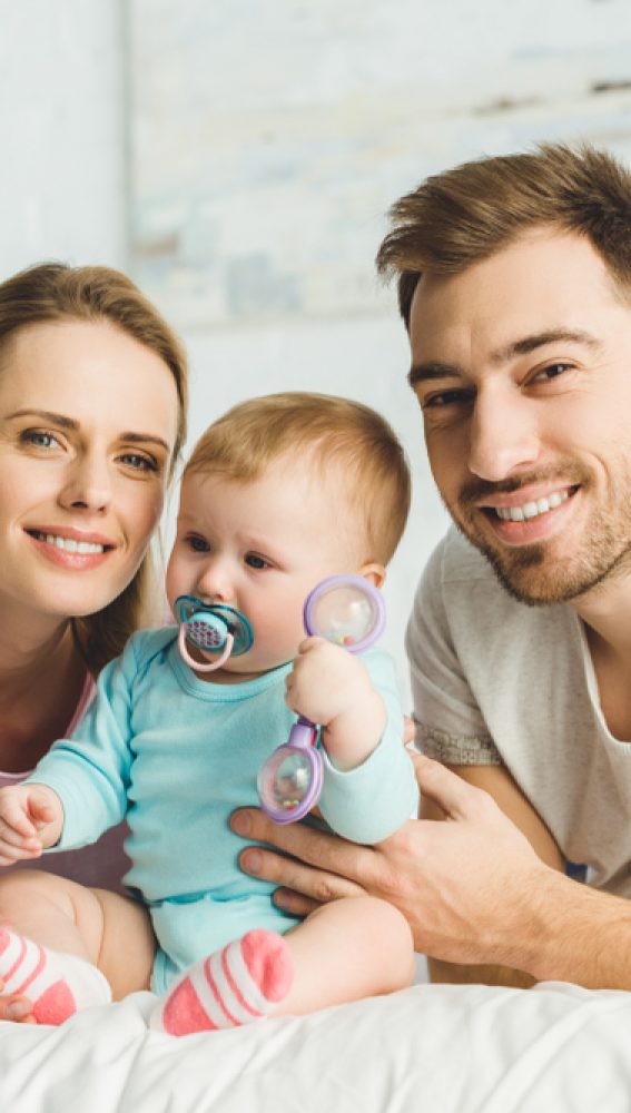 Smiling,Parents,Holding,Infant,Daughter,With,Baby,Dummy,And,Toy